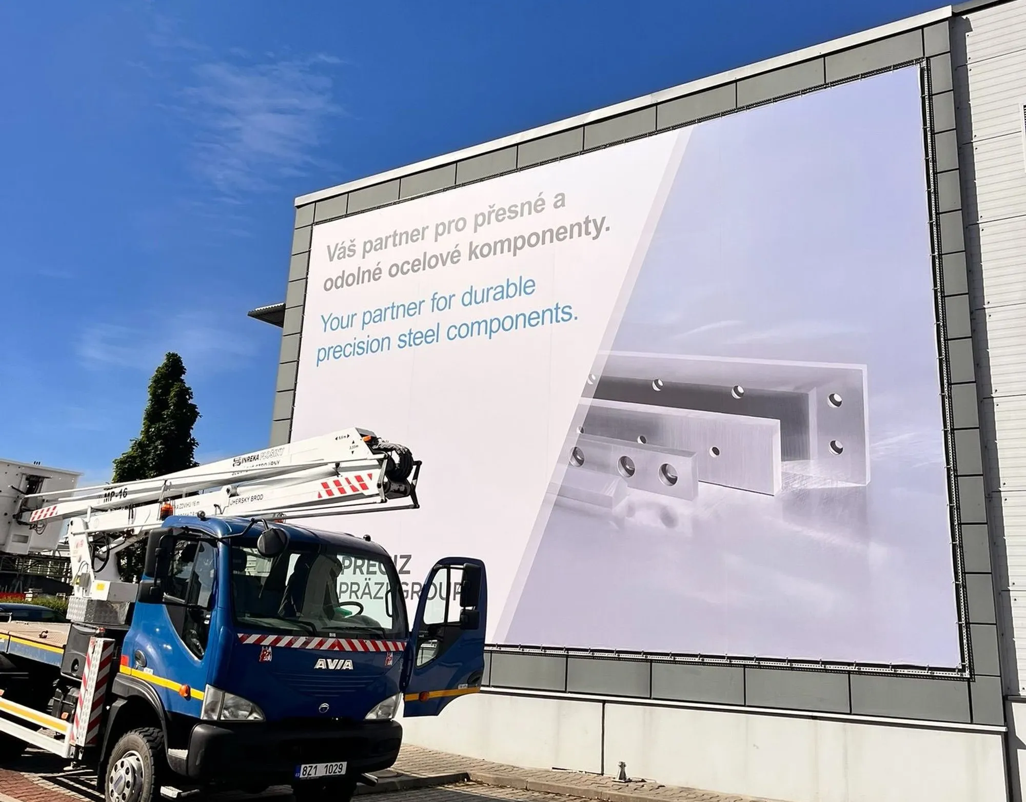 Large banner on the PRECIZ building with a slogan in Czech and English and a photo of steel parts, a lift truck parked in front